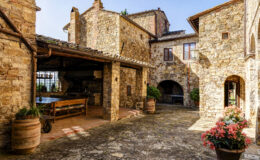 Charming stone courtyard of a rustic villa featuring a wooden dining table, potted plants, and vibrant flowers, bathed in warm sunlight.