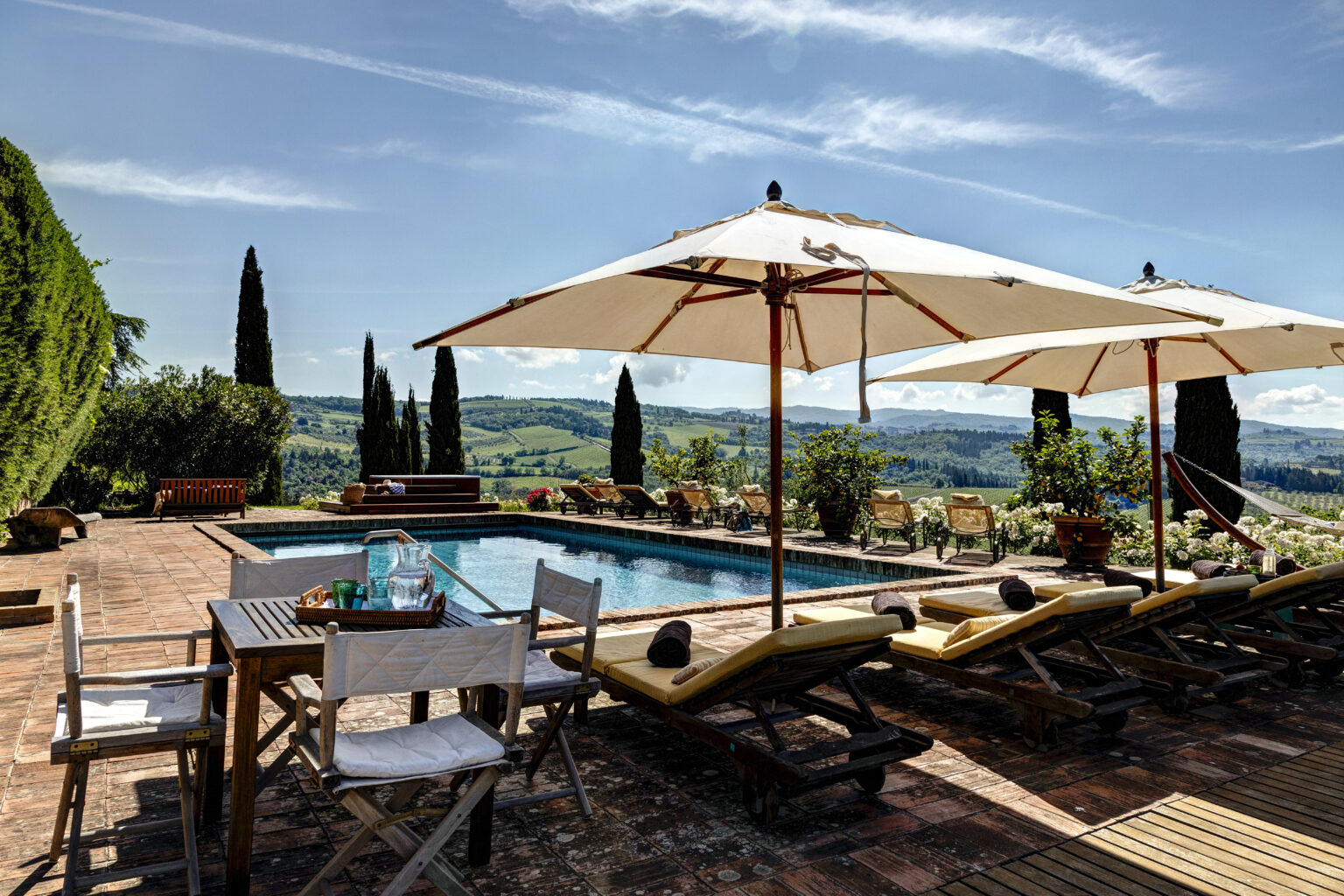 Scenic outdoor pool area with lounge chairs and umbrellas, surrounded by lush greenery and rolling hills, perfect for relaxation and leisure in a sunny setting. | Only on SopranoVillas