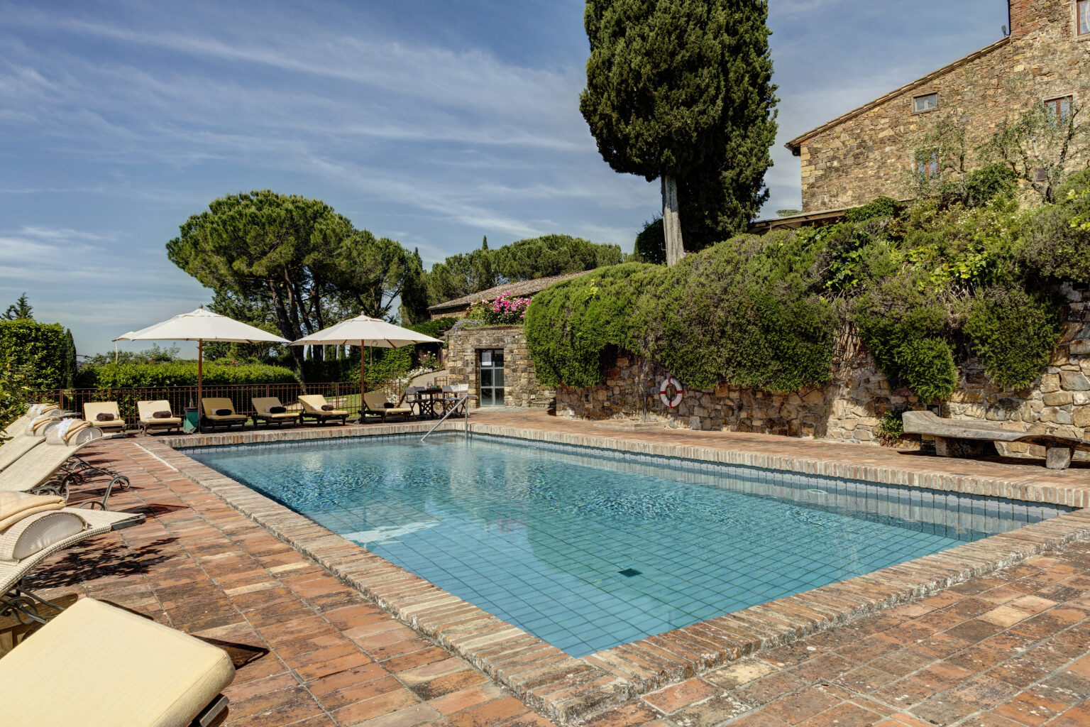 Luxurious outdoor swimming pool surrounded by sun loungers and umbrellas, set in a scenic garden with lush greenery and a rustic stone building in the background, perfect for relaxation and leisure. | Only on SopranoVillas