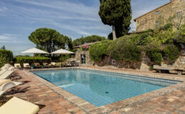 Luxurious outdoor swimming pool surrounded by sun loungers and umbrellas, set in a scenic garden with lush greenery and a rustic stone building in the background, perfect for relaxation and leisure.