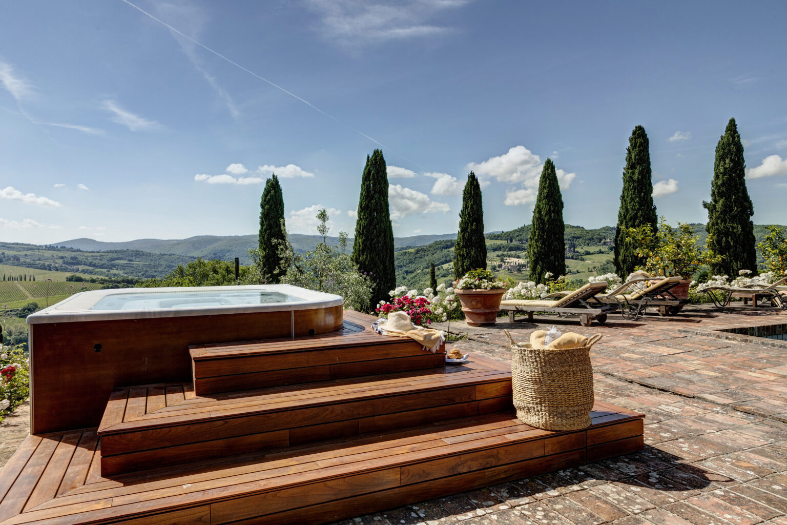 Luxurious outdoor spa area with wooden steps, surrounded by lush greenery and blooming flowers, overlooking rolling hills and a clear blue sky in a tranquil setting. | Only on SopranoVillas