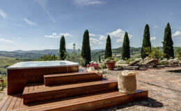 Luxurious outdoor spa area with wooden steps, surrounded by lush greenery and blooming flowers, overlooking rolling hills and a clear blue sky in a tranquil setting.