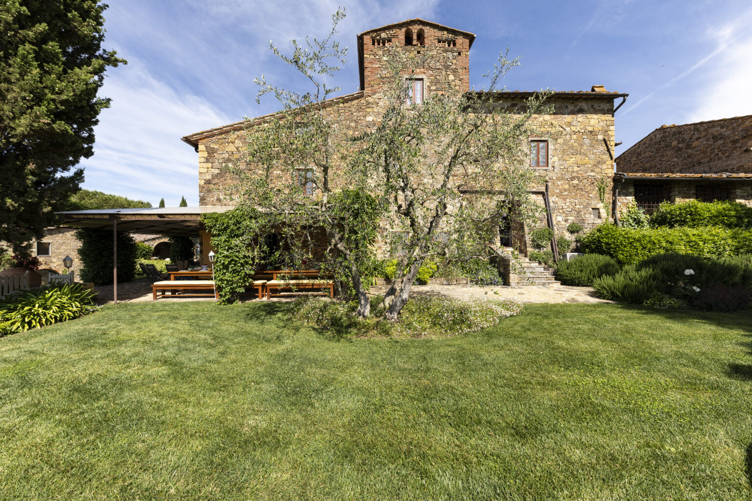 Stone farmhouse surrounded by lush green lawn and garden, featuring a large olive tree and outdoor seating area under a pergola. The structure showcases traditional architecture with a rustic charm, set against a clear blue sky. | Only on SopranoVillas