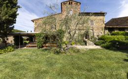 Stone farmhouse surrounded by lush green lawn and garden, featuring a large olive tree and outdoor seating area under a pergola. The structure showcases traditional architecture with a rustic charm, set against a clear blue sky.