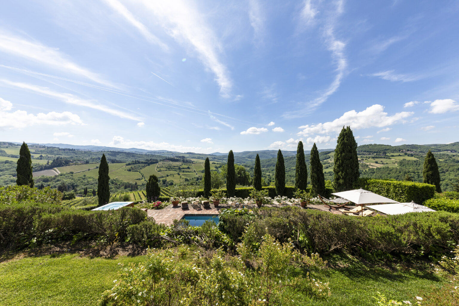 Scenic view of a luxurious villa in Tuscany featuring lush gardens, swimming pools, and cypress trees against a backdrop of rolling hills and blue skies. Ideal for travelers seeking relaxation and picturesque landscapes. | Only on SopranoVillas