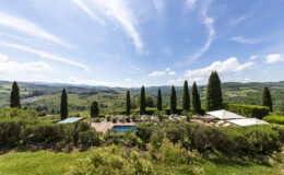 Scenic view of a luxurious villa in Tuscany featuring lush gardens, swimming pools, and cypress trees against a backdrop of rolling hills and blue skies. Ideal for travelers seeking relaxation and picturesque landscapes.