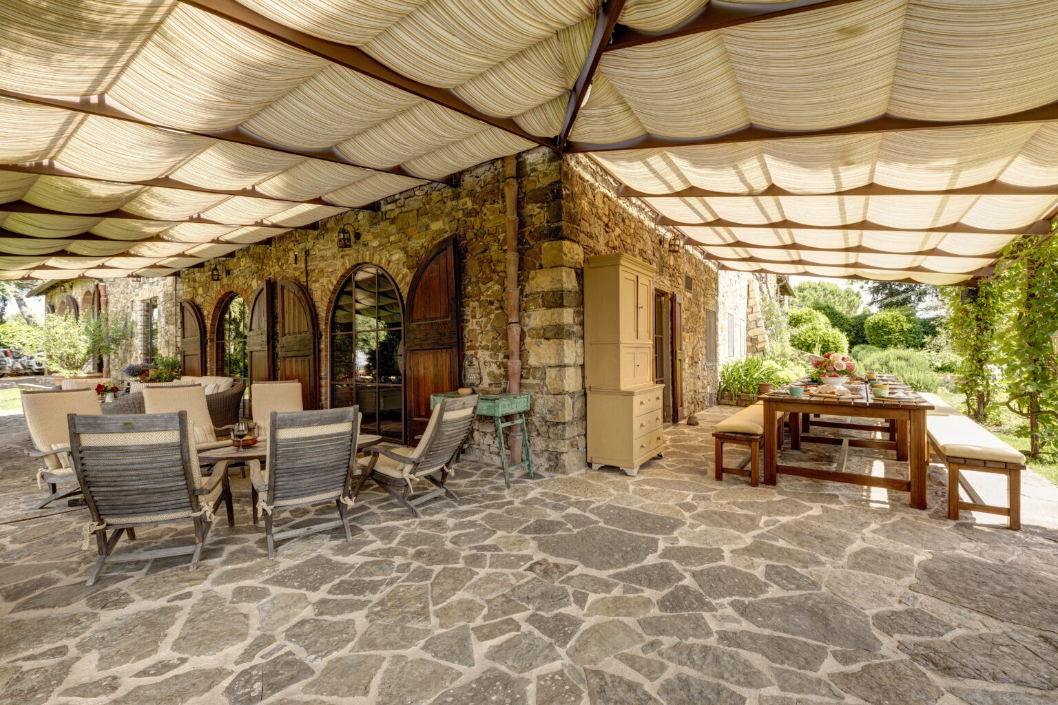 Outdoor dining area with stone flooring, featuring wooden furniture and a shaded patio. The space is adorned with plants and rustic stone walls, creating a cozy and inviting atmosphere for gatherings. | Only on SopranoVillas