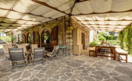 Outdoor dining area with stone flooring, featuring wooden furniture and a shaded patio. The space is adorned with plants and rustic stone walls, creating a cozy and inviting atmosphere for gatherings.