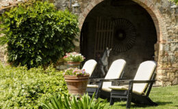 Cozy outdoor seating area featuring two lounge chairs with soft cushions, surrounded by lush greenery and colorful flower pots, set against a rustic stone wall with an arched entrance. A vintage horse statue is partially visible in the background, adding charm to the serene garden ambiance.