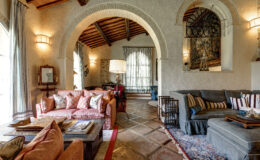 Cozy living room featuring a mix of vintage and modern decor, with a coral sofa adorned with decorative pillows, a wooden coffee table, and elegant lighting. The space showcases arched doorways, textured walls, and a warm color palette, creating an inviting atmosphere.
