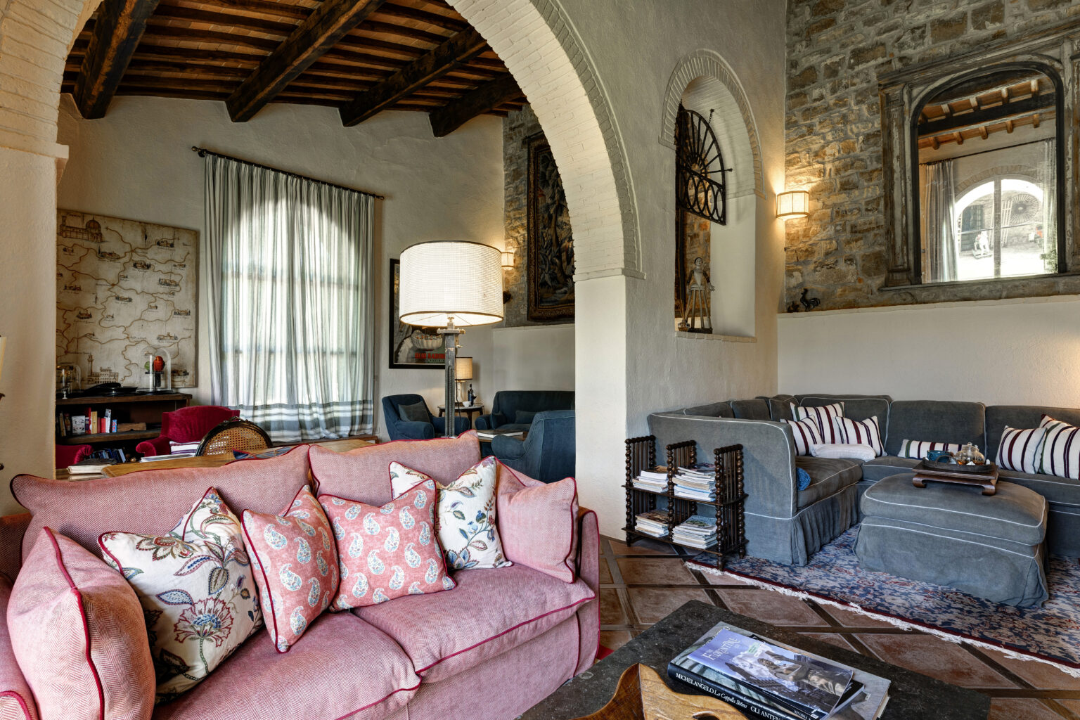 Cozy living room featuring a pink sofa adorned with decorative pillows, a stylish lamp, and a mix of modern and rustic decor elements, including a stone wall and wooden beams. The space is brightened by large windows with sheer curtains, creating an inviting atmosphere for relaxation. | Only on SopranoVillas