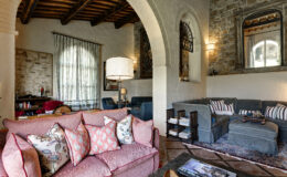 Cozy living room featuring a pink sofa adorned with decorative pillows, a stylish lamp, and a mix of modern and rustic decor elements, including a stone wall and wooden beams. The space is brightened by large windows with sheer curtains, creating an inviting atmosphere for relaxation.