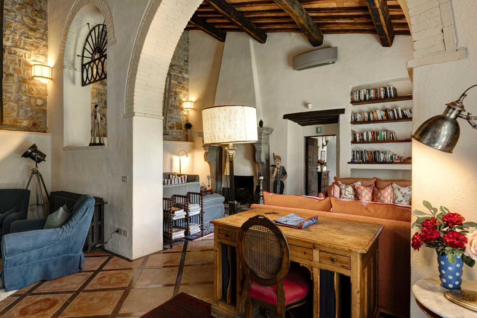 Cozy living room featuring a rustic wooden desk, colorful seating, and a bookshelf filled with books. The space is illuminated by stylish lamps and has stone walls, arched ceilings, and a warm ambiance, perfect for relaxation or reading. | Only on SopranoVillas