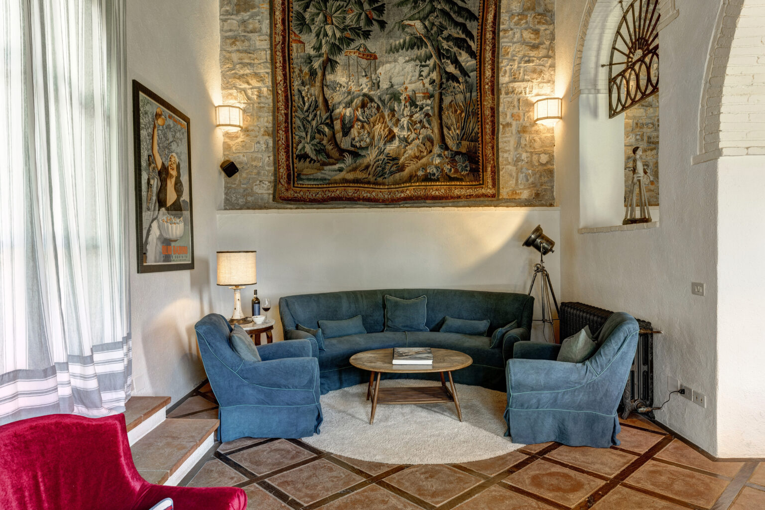 Cozy living room featuring a curved teal sofa and armchairs, a round wooden coffee table, and a decorative tapestry on the stone wall. Natural light filters through sheer curtains, complemented by stylish lighting fixtures and vintage decor elements. | Only on SopranoVillas