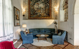 Cozy living room featuring a curved teal sofa and armchairs, a round wooden coffee table, and a decorative tapestry on the stone wall. Natural light filters through sheer curtains, complemented by stylish lighting fixtures and vintage decor elements.