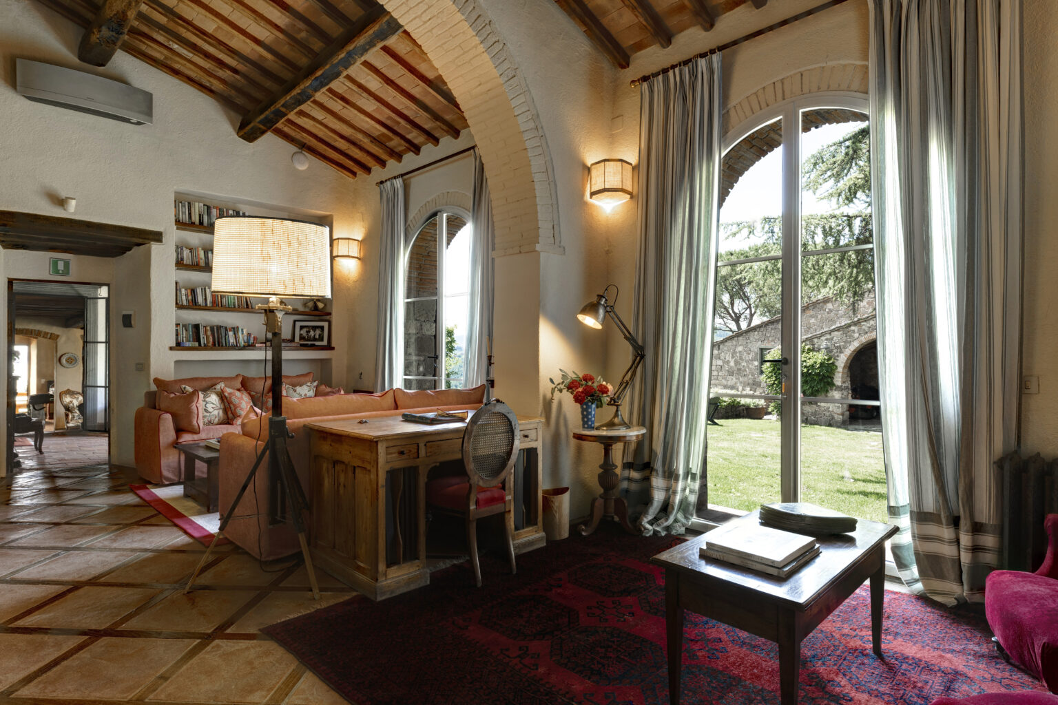 Cozy living room interior featuring a rustic wooden desk, comfortable sofa with decorative pillows, and large windows with sheer curtains. The space is enhanced by warm lighting and a view of the garden outside, creating an inviting atmosphere perfect for relaxation or work. | Only on SopranoVillas