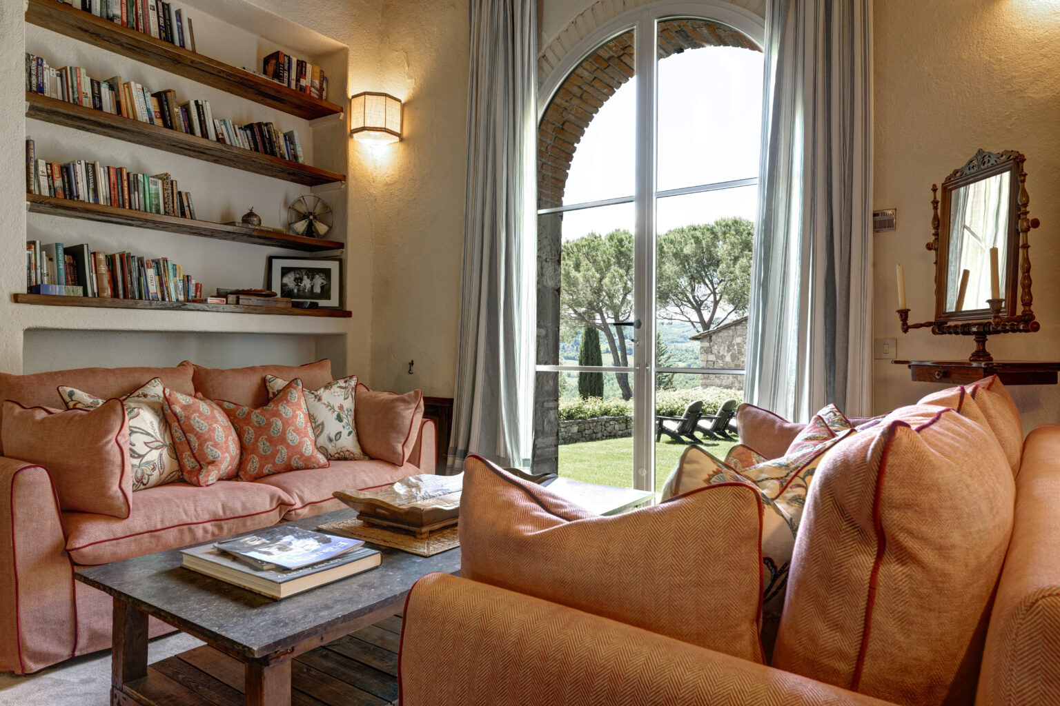 Cozy living room featuring a comfortable coral sofa adorned with patterned cushions, a rustic wooden coffee table with books, and a wall shelf filled with books. Large arched windows offer a view of a landscaped garden with trees, enhancing the inviting atmosphere. | Only on SopranoVillas
