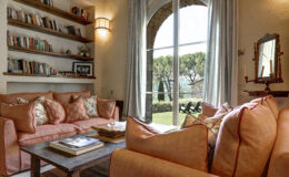 Cozy living room featuring a comfortable coral sofa adorned with patterned cushions, a rustic wooden coffee table with books, and a wall shelf filled with books. Large arched windows offer a view of a landscaped garden with trees, enhancing the inviting atmosphere.