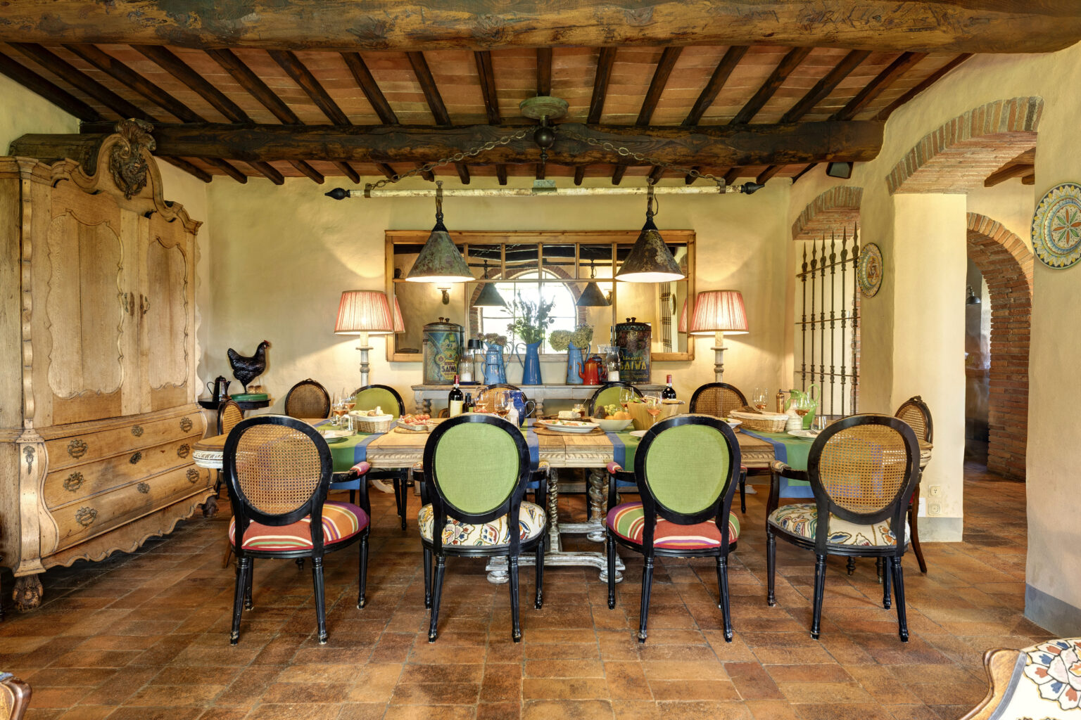 Rustic dining room featuring a large wooden table set for a meal, surrounded by colorful chairs. The room has exposed wooden beams, vintage lighting fixtures, and decorative elements like a wooden armoire and a rooster statue, creating a warm and inviting atmosphere. | Only on SopranoVillas