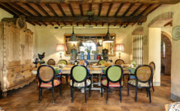 Rustic dining room featuring a large wooden table set for a meal, surrounded by colorful chairs. The room has exposed wooden beams, vintage lighting fixtures, and decorative elements like a wooden armoire and a rooster statue, creating a warm and inviting atmosphere.