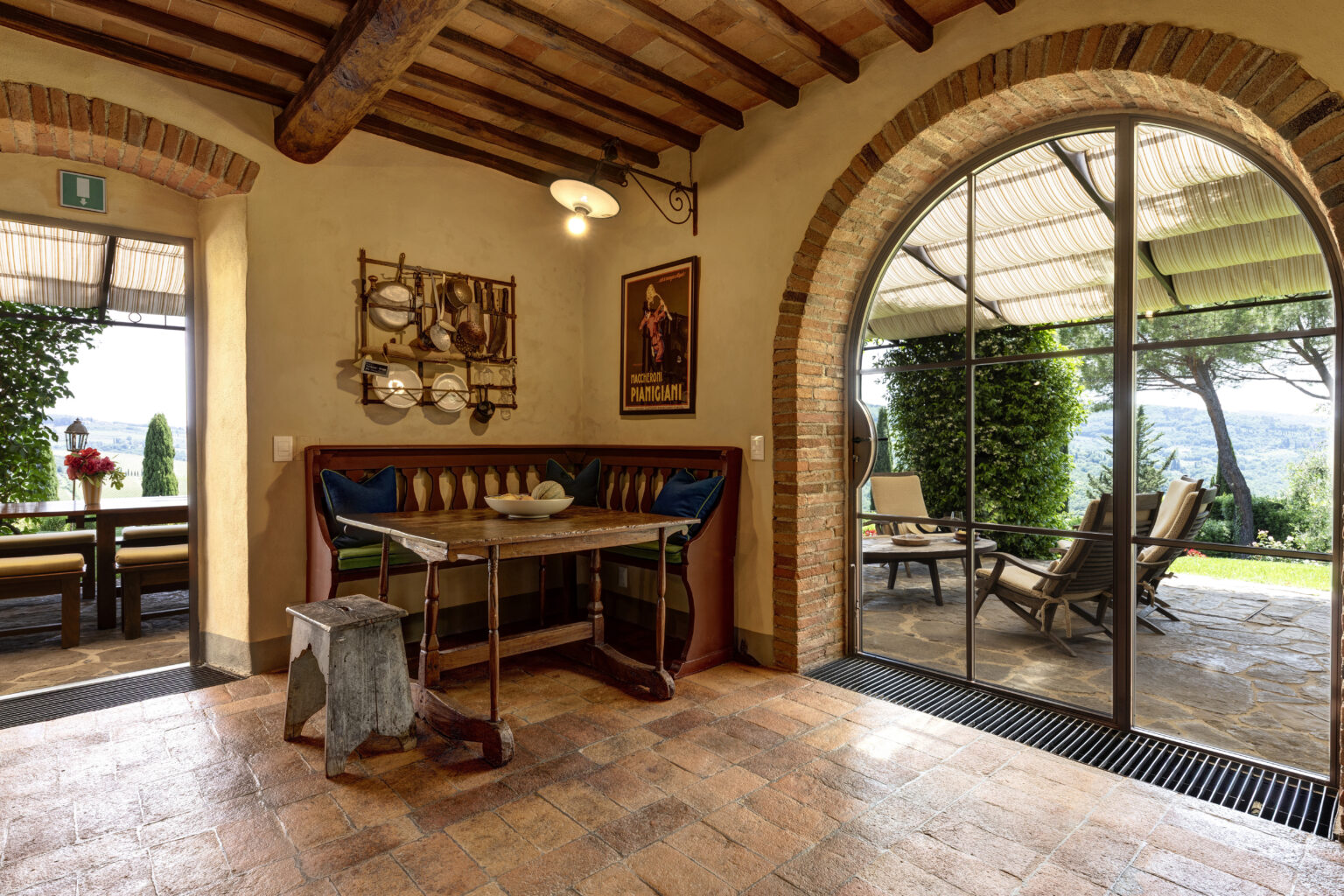 Cozy dining area with rustic wooden table and bench seating, featuring a decorative wall rack with kitchen utensils. An arched window reveals a scenic outdoor patio with comfortable seating, surrounded by greenery and picturesque views. Warm, earthy tones and terracotta flooring enhance the inviting atmosphere. | Only on SopranoVillas