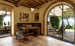Cozy dining area with rustic wooden table and bench seating, featuring a decorative wall rack with kitchen utensils. An arched window reveals a scenic outdoor patio with comfortable seating, surrounded by greenery and picturesque views. Warm, earthy tones and terracotta flooring enhance the inviting atmosphere.