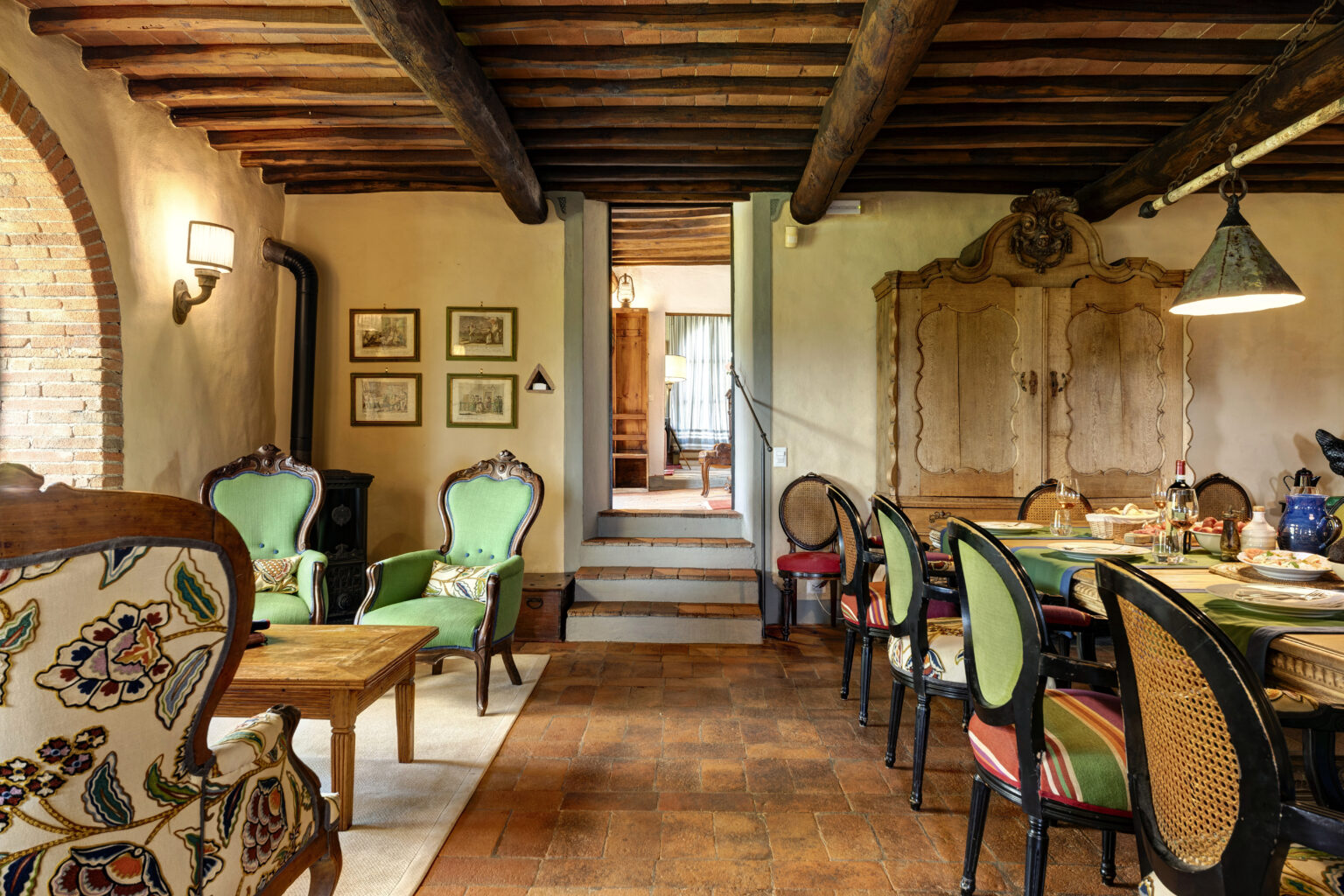 Cozy interior of a rustic dining and living area featuring vintage furniture, wooden beams, and warm terracotta flooring. The space includes green upholstered chairs, a wooden dining table set for a meal, and decorative wall art, creating an inviting atmosphere. | Only on SopranoVillas