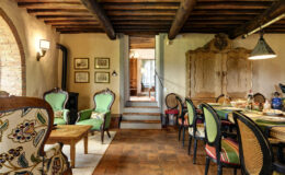 Cozy interior of a rustic dining and living area featuring vintage furniture, wooden beams, and warm terracotta flooring. The space includes green upholstered chairs, a wooden dining table set for a meal, and decorative wall art, creating an inviting atmosphere.