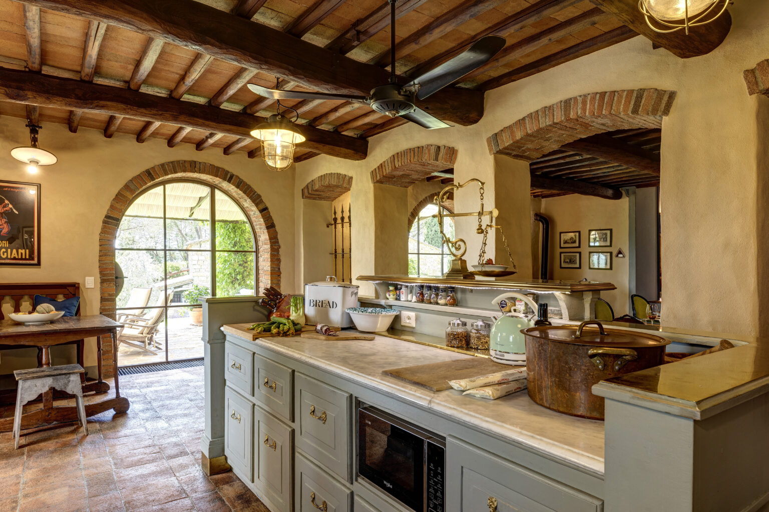 Rustic kitchen interior featuring a marble countertop, wooden cabinetry, and brick arches. The space includes a ceiling fan, decorative lighting, and a cozy dining area with a wooden table. Natural light floods in through large windows, highlighting kitchenware and fresh produce on the counter. | Only on SopranoVillas