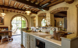Rustic kitchen interior featuring a marble countertop, wooden cabinetry, and brick arches. The space includes a ceiling fan, decorative lighting, and a cozy dining area with a wooden table. Natural light floods in through large windows, highlighting kitchenware and fresh produce on the counter.