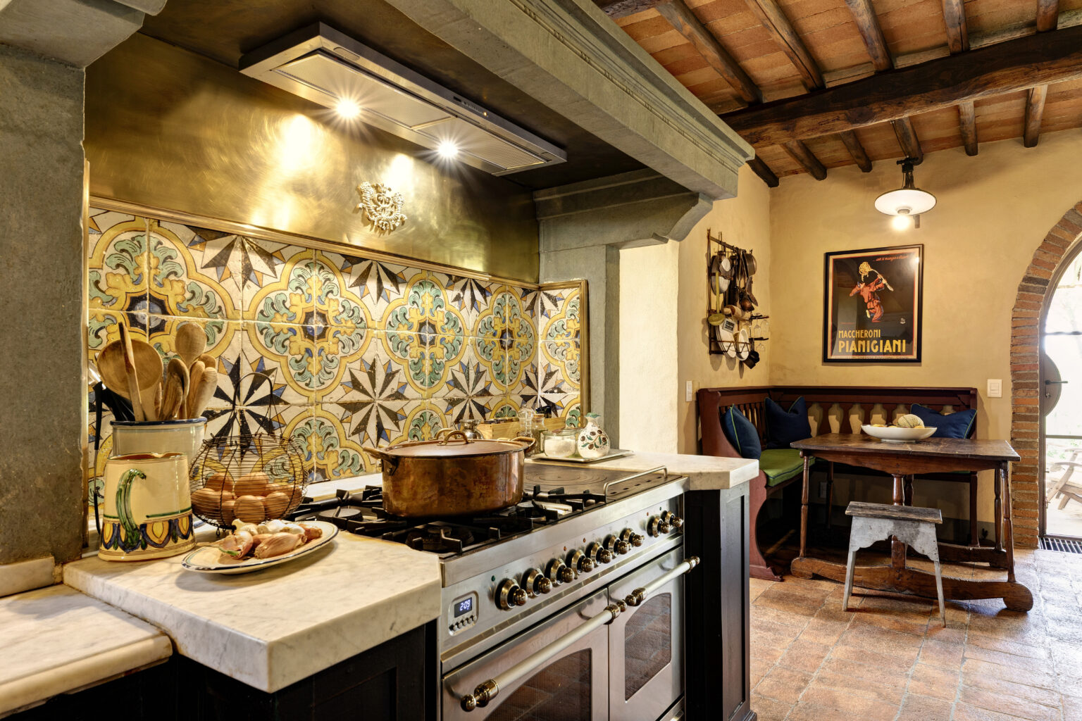 Rustic kitchen featuring a traditional gas stove, marble countertop, and colorful tiled backsplash. The space includes wooden utensils, a decorative pot, and a cozy dining area with a round wooden table and cushioned seating. Exposed wooden beams and warm lighting enhance the inviting atmosphere. | Only on SopranoVillas