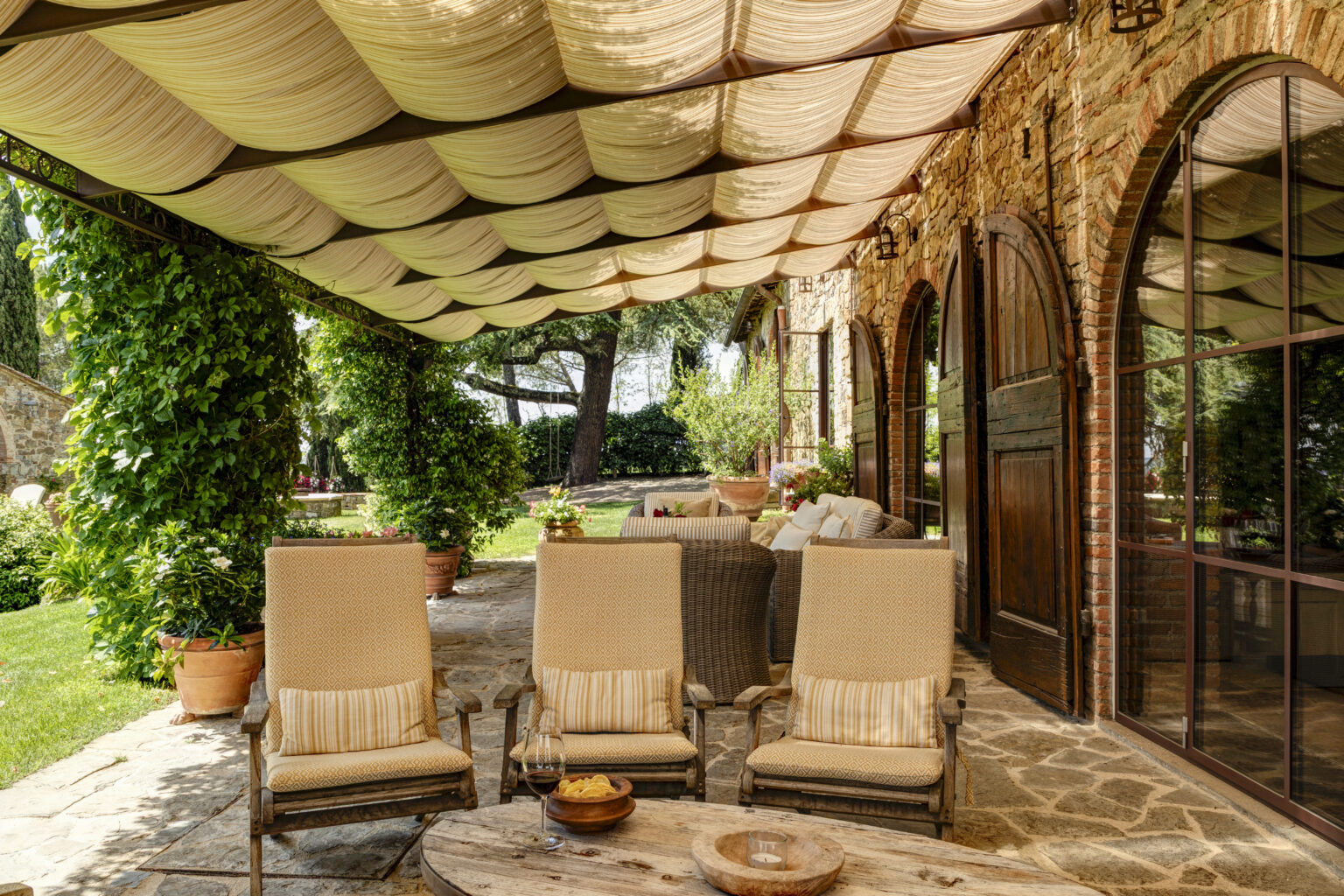 Cozy outdoor patio featuring four comfortable lounge chairs with striped cushions, a rustic wooden table with snacks and wine, and a beautifully draped canopy. Lush greenery and stone walls create a serene atmosphere, perfect for relaxation. | Only on SopranoVillas