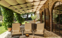Cozy outdoor patio featuring four comfortable lounge chairs with striped cushions, a rustic wooden table with snacks and wine, and a beautifully draped canopy. Lush greenery and stone walls create a serene atmosphere, perfect for relaxation.