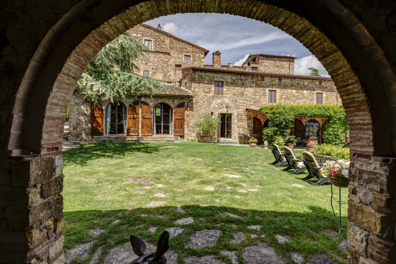 View of a charming Italian villa courtyard framed by an archway, featuring lush green grass, stone pathways, and wooden doors leading to the main building. The scene includes comfortable outdoor seating and vibrant flower pots, showcasing a serene and inviting atmosphere. | Only on SopranoVillas