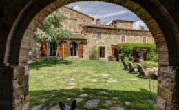 View of a charming Italian villa courtyard framed by an archway, featuring lush green grass, stone pathways, and wooden doors leading to the main building. The scene includes comfortable outdoor seating and vibrant flower pots, showcasing a serene and inviting atmosphere.