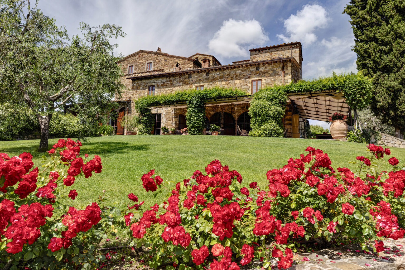 Charming stone villa surrounded by lush greenery and vibrant red roses, set against a bright blue sky with fluffy clouds, ideal for a picturesque getaway in the countryside. | Only on SopranoVillas