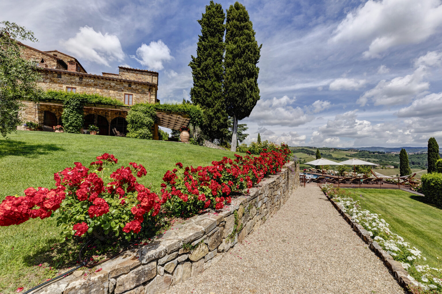 Scenic view of a Tuscan villa surrounded by vibrant red flowers, lush green lawns, and a backdrop of rolling hills under a blue sky with clouds. The stone pathway leads to a cozy outdoor seating area with umbrellas, showcasing the beauty of the Italian countryside. | Only on SopranoVillas