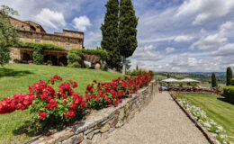 Scenic view of a Tuscan villa surrounded by vibrant red flowers, lush green lawns, and a backdrop of rolling hills under a blue sky with clouds. The stone pathway leads to a cozy outdoor seating area with umbrellas, showcasing the beauty of the Italian countryside.