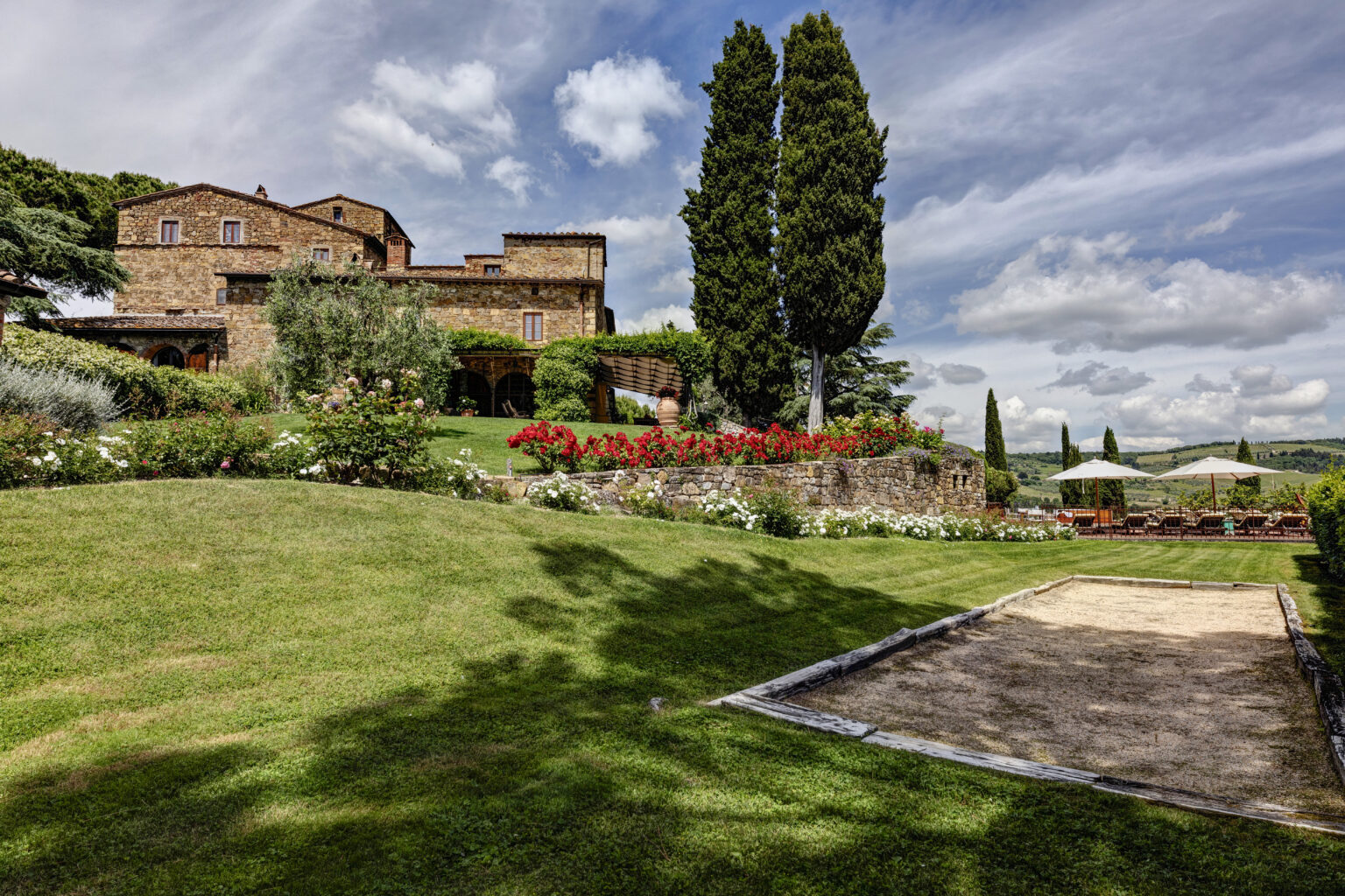 Scenic view of a rustic stone villa surrounded by lush gardens and vibrant flowers, featuring outdoor seating areas under umbrellas, with rolling hills in the background and a clear blue sky. Ideal for vacation rentals in Tuscany. | Only on SopranoVillas