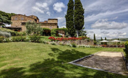 Scenic view of a rustic stone villa surrounded by lush gardens and vibrant flowers, featuring outdoor seating areas under umbrellas, with rolling hills in the background and a clear blue sky. Ideal for vacation rentals in Tuscany.