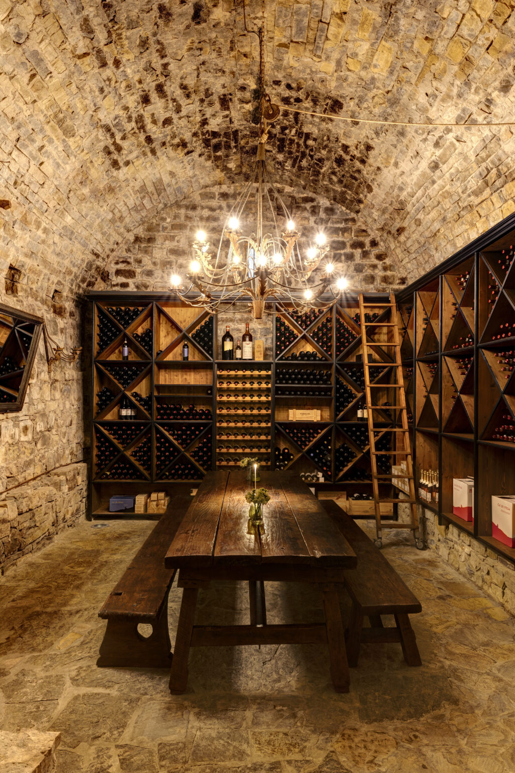 Wine cellar interior featuring rustic stone walls, wooden wine racks filled with bottles, a large wooden table adorned with a small vase of flowers, and a chandelier providing warm lighting. | Only on SopranoVillas