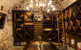 Wine cellar interior featuring rustic stone walls, wooden wine racks filled with bottles, a large wooden table adorned with a small vase of flowers, and a chandelier providing warm lighting.