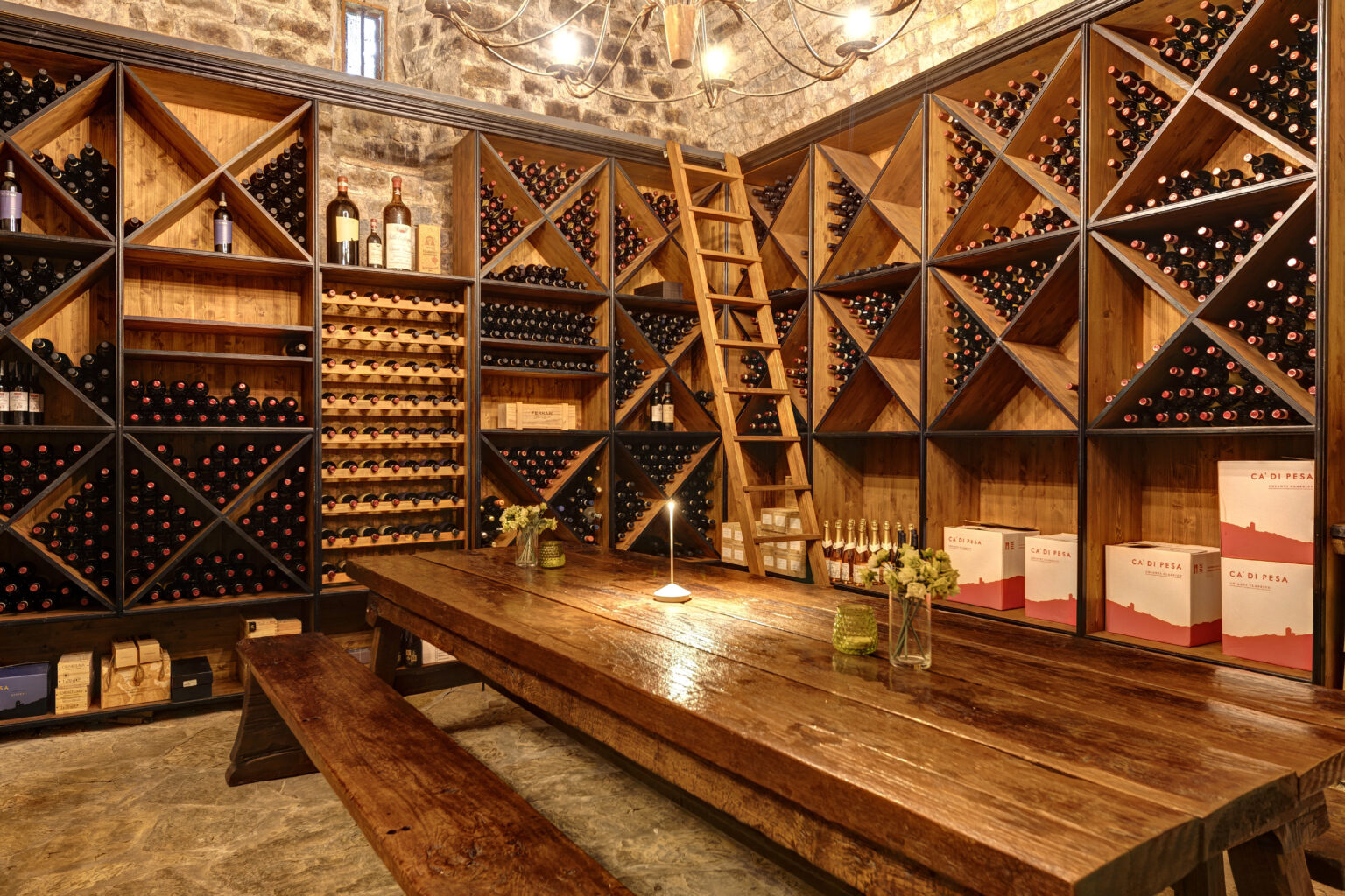 Wine cellar interior featuring wooden shelves filled with various wine bottles, a rustic wooden table, and a ladder for accessing upper shelves, illuminated by warm lighting. | Only on SopranoVillas