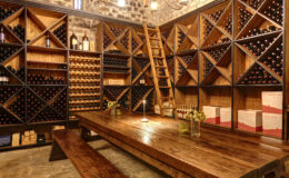 Wine cellar interior featuring wooden shelves filled with various wine bottles, a rustic wooden table, and a ladder for accessing upper shelves, illuminated by warm lighting.