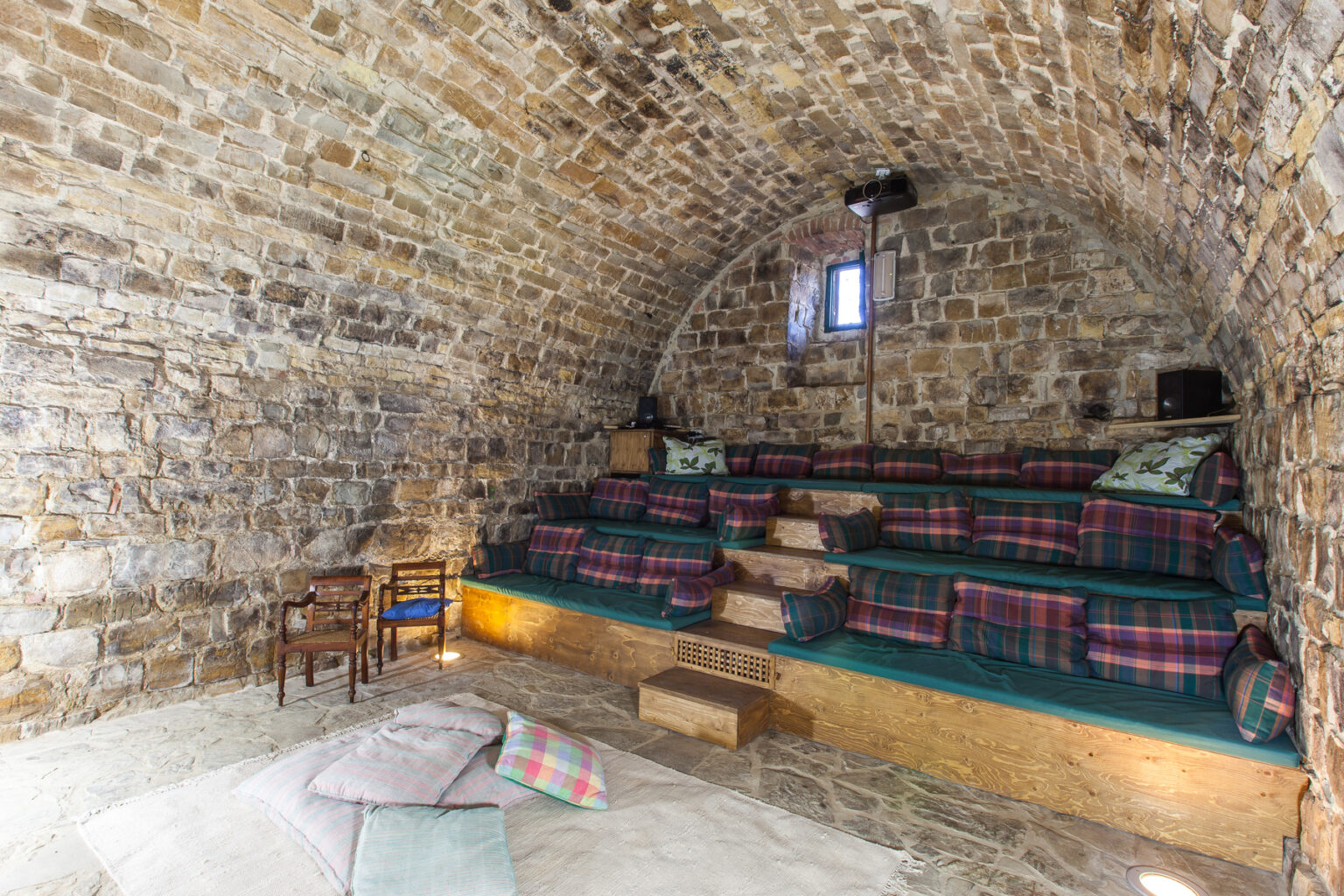 Cozy interior of a stone-walled room featuring tiered seating with plaid cushions, wooden accents, and a soft rug, ideal for relaxation or gatherings. | Only on SopranoVillas