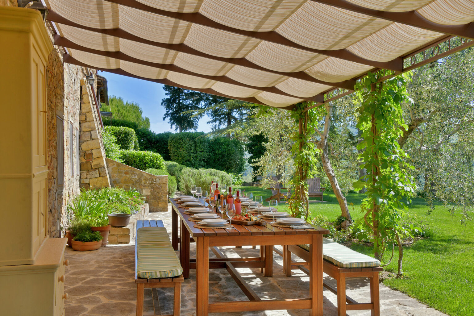 Outdoor dining area under a pergola, featuring a long wooden table set for a meal, surrounded by lush greenery and stone walls, ideal for al fresco dining and gatherings. | Only on SopranoVillas
