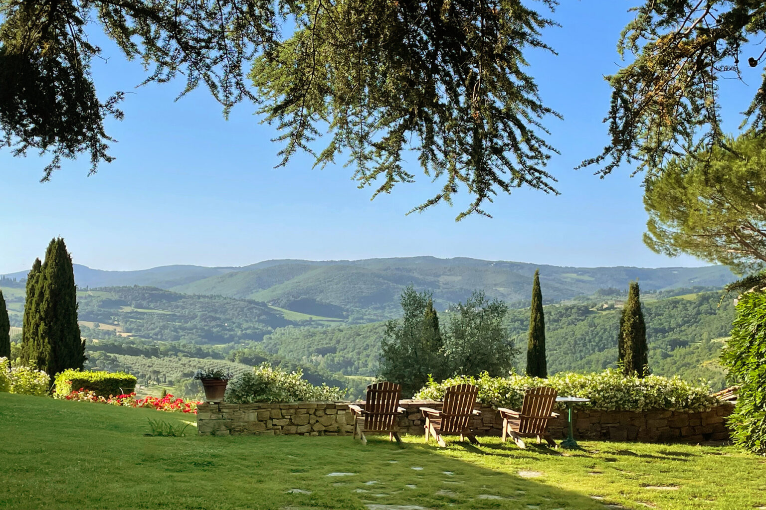 Scenic view of a lush green landscape with rolling hills and a clear blue sky, featuring a cozy seating area with wicker chairs and vibrant flower beds in the foreground. Ideal for nature lovers and outdoor enthusiasts. | Only on SopranoVillas