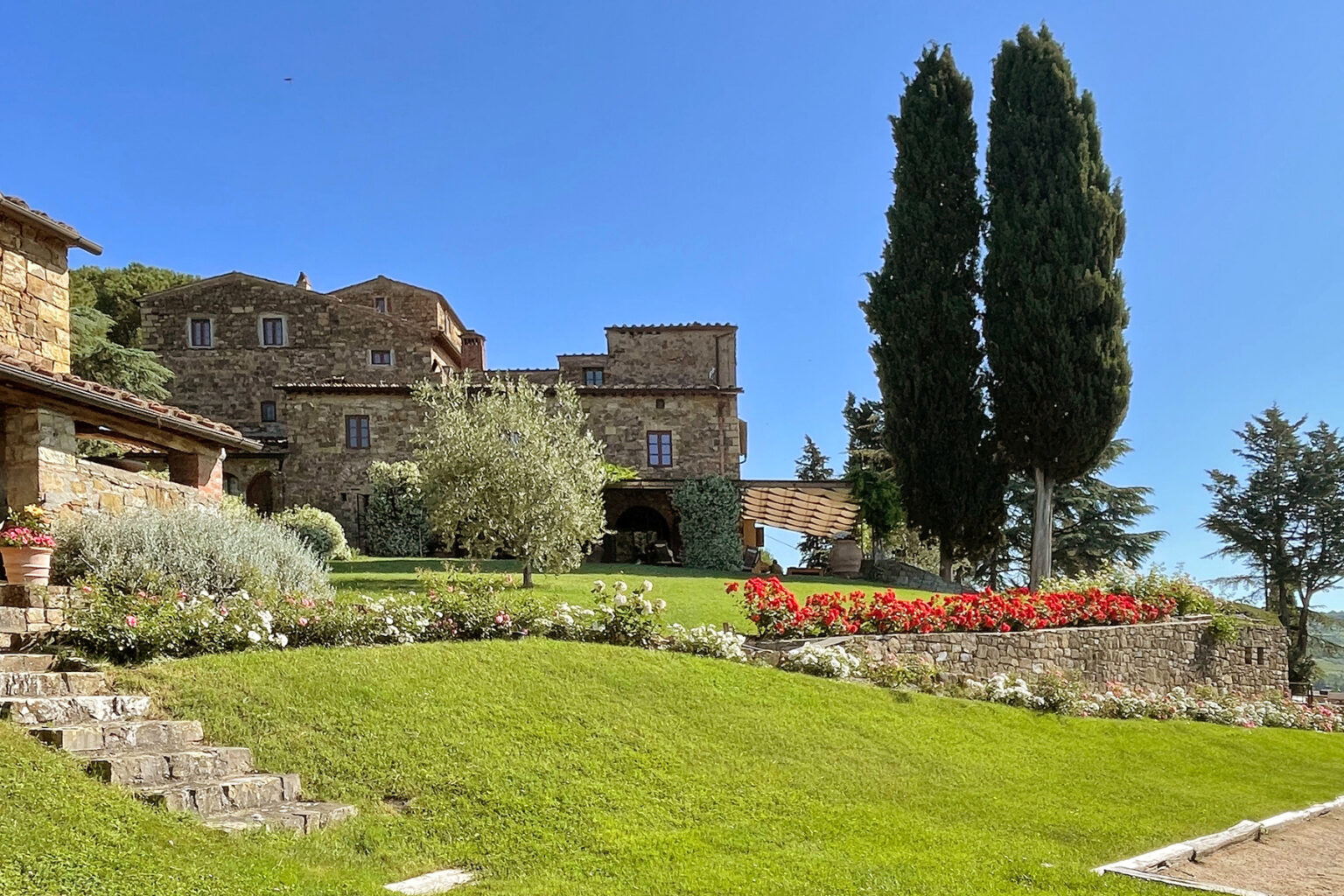 Scenic view of a rustic stone villa surrounded by lush green lawns, blooming flowers, and tall cypress trees under a clear blue sky. | Only on SopranoVillas