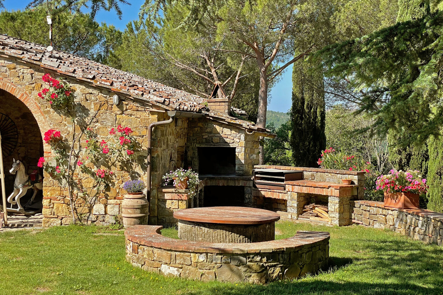 Rustic outdoor patio with stone barbecue area, surrounded by vibrant flowers and lush greenery, featuring a charming stone structure under a clear blue sky. | Only on SopranoVillas