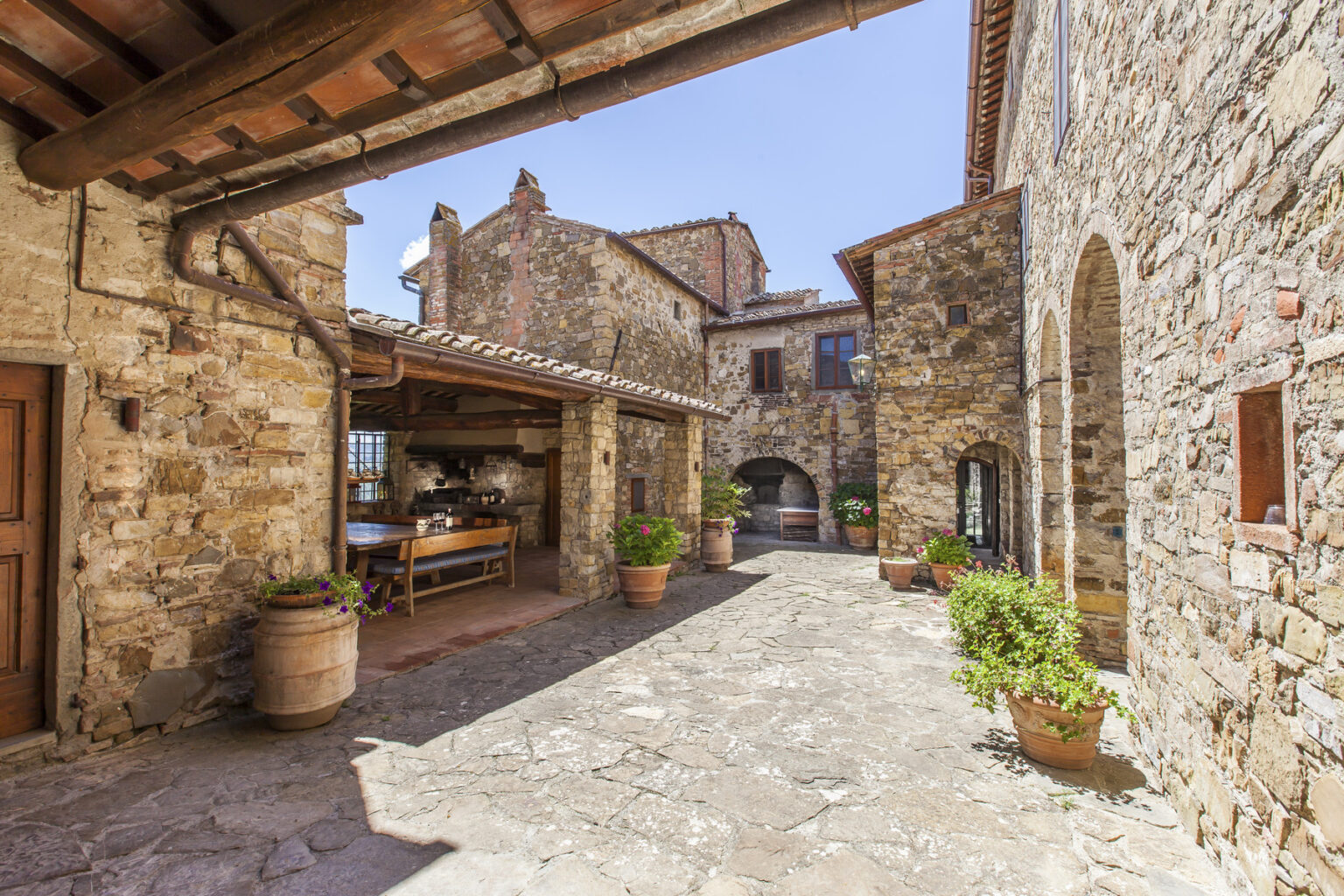 Rustic stone courtyard featuring traditional Tuscan architecture, with terracotta pots and a wooden dining table under a covered area, surrounded by historic buildings and blue sky. | Only on SopranoVillas